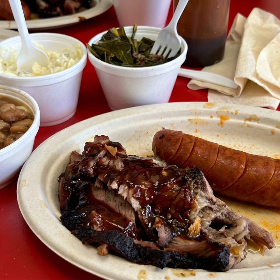 Barbecue plate with sliced sausage and smoked meat, accompanied by beans, creamy side dish, greens, bread, and drinks on a red table.