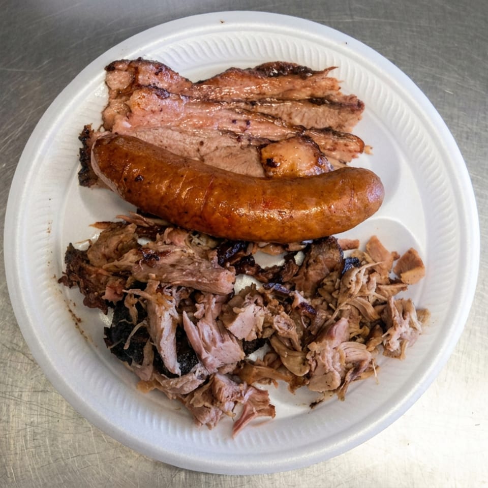 Plate of grilled sausage, sliced roasted meat, and shredded barbecue pork on a white disposable plate.