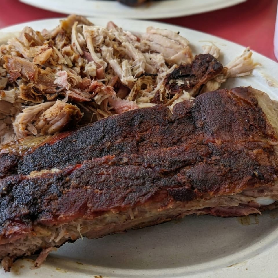 Plate of smoked barbecue ribs and shredded meat on a disposable plate, with a drink and additional ribs nearby.